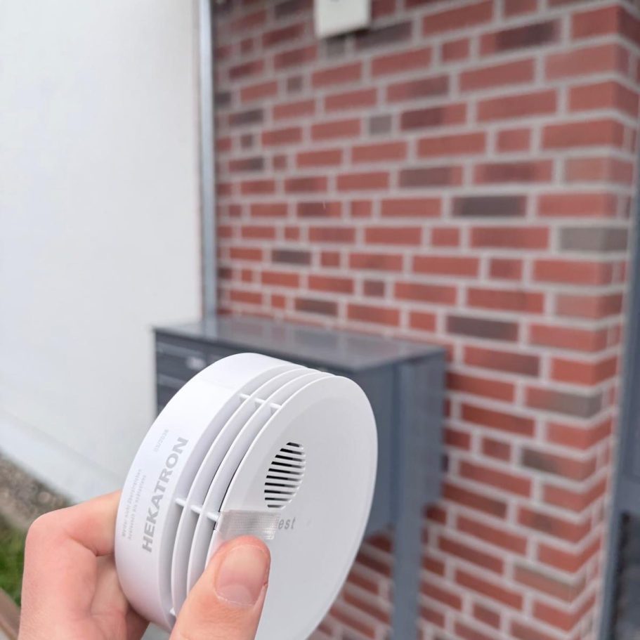 Person hält Rauchmelder vor einer Hauswand in der Hand