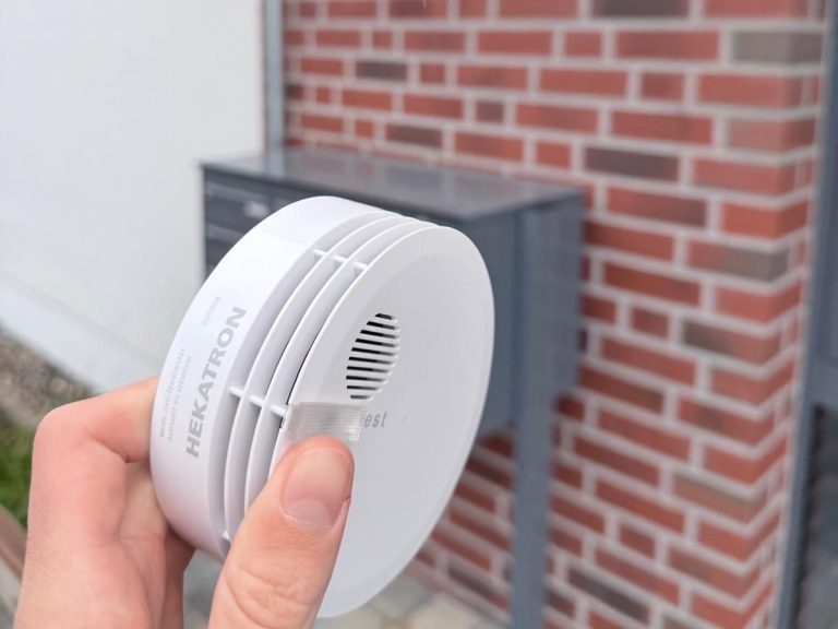 Person hält Rauchmelder vor einer Hauswand in der Hand